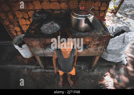 Enfant atteint de pauvreté debout devant une cuisine improvisée à l'extérieur d'une maison de boue Banque D'Images