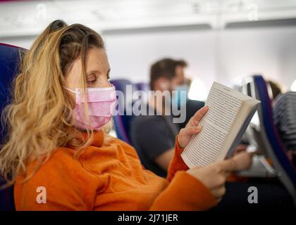 Femme mature qui lit dans l'avion en cas de pandémie avec un masque rose portant de l'orange et des passagers portant un masque en arrière-plan Banque D'Images