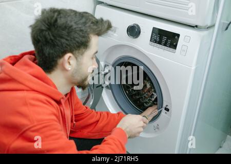 Jeune homme portant un sweat à capuche rouge fixant le lave-linge dans la salle de bains, gros plan. Banque D'Images