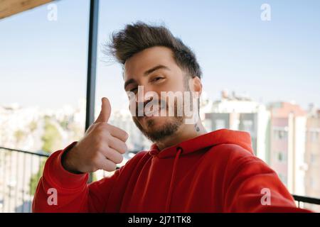 Beau jeune homme portant un sweat à capuche rouge prenant un autoportrait à l'extérieur sur le balcon. Un homme heureux souriant sur l'appareil photo avec le pouce vers le haut tout en prenant le selfie. Banque D'Images