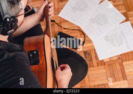 vue de dessus, homme assis sur le sol étudiant la musique avec guitare acoustique, casque, partitions et un téléphone sur le sol. Banque D'Images