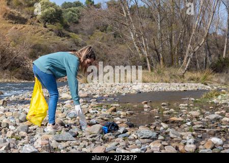 Une femme qui s'accroupe vers le bas pour ramasser une bouteille en plastique trouvée parmi les pierres de rivière. Concept d'écologie Banque D'Images