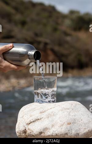 Gros plan versant de l'eau de boisson fraîche purifiée de la bouteille sur une tasse en verre dans la nature avec la rivière derrière Banque D'Images