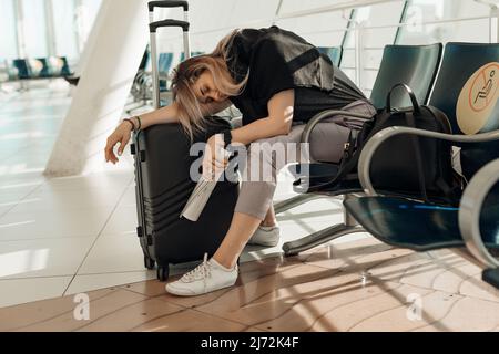 Femme blonde fatiguée et somnolente vêtue de vêtements décontractés avec bagages, penchée sur des sacs, assise dans le terminal de l'aéroport. Annuler le vol Banque D'Images