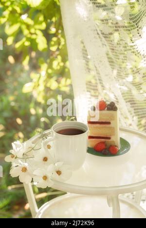 Belle tasse de café en porcelaine et une tranche de gâteau sur table en verre décorée de fleurs blanches dans le jardin d'été à la lumière du soleil.Copier l'espace.Concept des boissons d'été. Banque D'Images