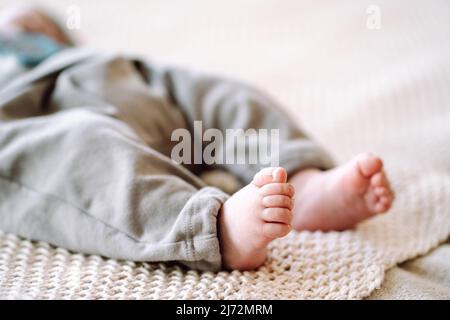 Photo courte du nouveau-né pieds nus portant un body gris couché sur un tissu tricoté en coton beige. Gros plan des adorables petits pieds de bébé sur le lit. Literie Banque D'Images