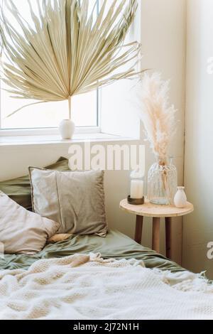 Intérieur de chambre moderne ensoleillée avec lit recouvert de linge de lit gris, écossais beige. Grand vase en verre avec pampas sec, bougie décorative sur table de chevet Banque D'Images