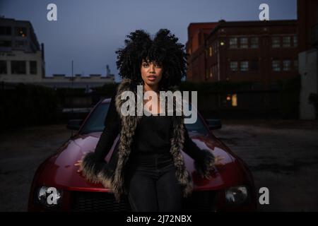 Jeune femme afro-américaine confiante assise sur le capot de sa voiture et regardant dans la caméra. Banque D'Images