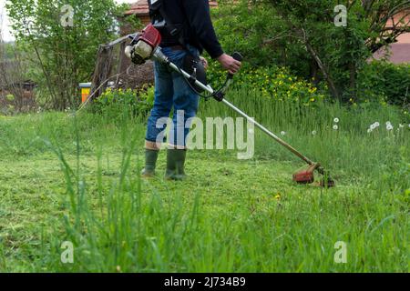 Homme fauchant de l'herbe haute avec un coupe-herbe à essence dans la cour d'une maison de campagne. Équipement de jardinage. Processus de tonte de la pelouse avec la tondeuse à main Banque D'Images