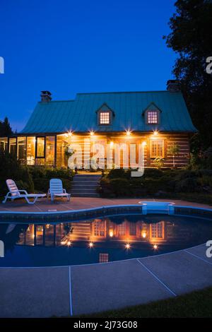 Vue arrière de l'ancienne maison en rondins de style Canadiana à deux étages datant de 1800s avec jardin paysagé et piscine au crépuscule. Banque D'Images