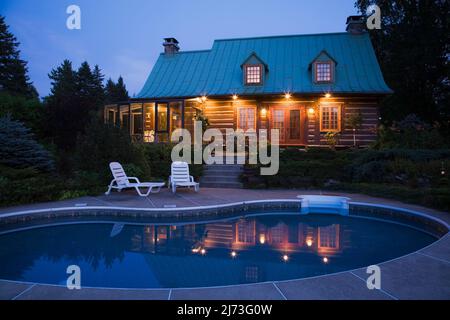 Vue arrière de l'ancienne maison en rondins de style Canadiana à deux étages datant de 1800s avec jardin paysagé et piscine au crépuscule. Banque D'Images