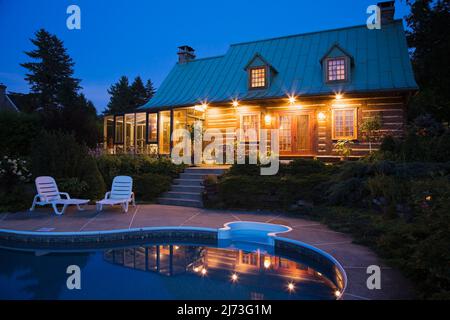 Vue arrière de l'ancienne maison en rondins de style Canadiana à deux étages datant de 1800s avec jardin paysagé et piscine au crépuscule. Banque D'Images