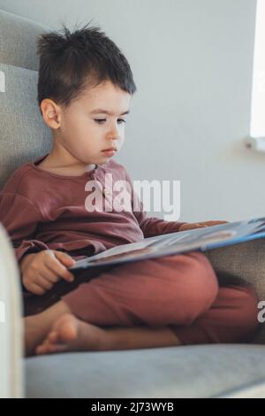 Un garçon d'âge scolaire lisant un livre tout en étant à l'aise sur le canapé pour faire ses devoirs à l'école. Enseignement à distance. Gros plan. Banque D'Images