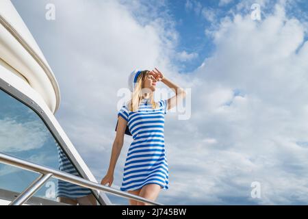 Une femme navigueuse se tient sur un pont de yacht avec une robe rayée et un chapeau regarde dans la distance par rapport à l'arrière-plan d'un ciel bleu avec des nuages. Une fille caucasienne aime ses vacances sur un yacht Banque D'Images