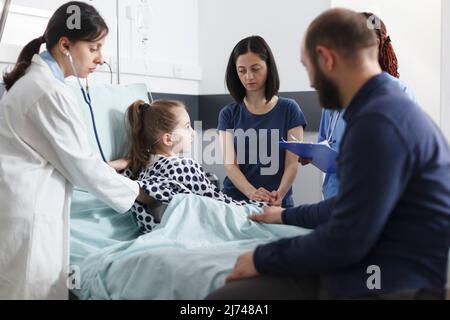 Pédiatre consultant malade petite fille en utilisant stéthoscope tout en inquiet parents attendant en plus d'elle. Médecin généraliste examinant une fille malade dans la salle de service des patients de la clinique pédiatrique. Banque D'Images