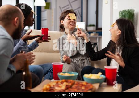 Groupe diversifié de collègues jouant deviner qui jeu avec des notes adhésives sur le front, appréciant l'activité amusante avec des charades jouer. Les gens devinrent le pantomime à la fête des boissons après les heures de travail. Banque D'Images