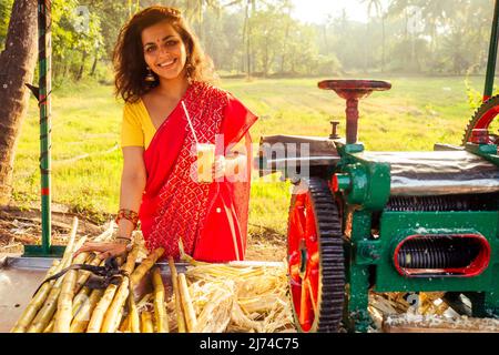 belle femme induan en rouge sari debout près de la machine à jus de canne à sucre appareil machine , plantation d'été ferme arrière-plan. petite entreprise de démarrage Banque D'Images