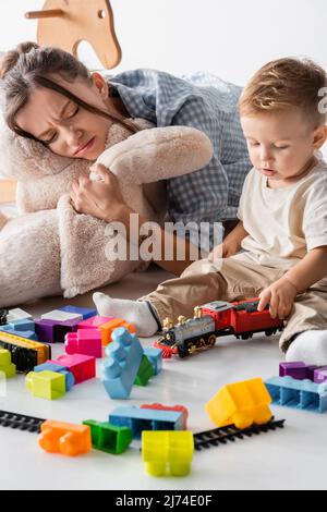 une femme qui frogne s'enserre un jouet doux près d'un enfant en bas âge jouant sur du blanc Banque D'Images