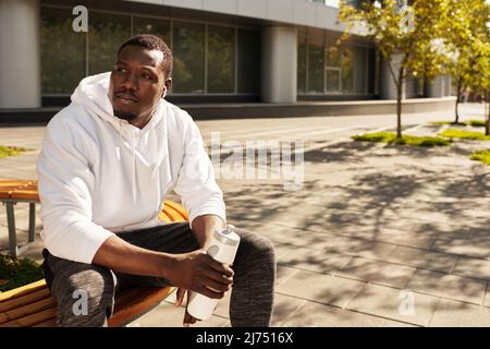 Jeune coureur afro-américain se reposant après l'entraînement à l'extérieur, assis sur le banc avec une bouteille d'eau, en écoutant de la musique à l'aide d'écouteurs Banque D'Images