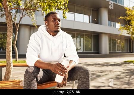 Jeune athlète masculin noir se détendant après l'entraînement de course, assis sur le banc avec une bouteille d'eau à l'extérieur, contemplant et appréciant le matin ensoleillé Banque D'Images