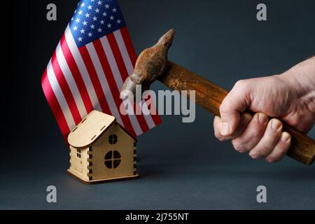 Un marteau est élevé au-dessus de la maison avec le drapeau des Etats-Unis. Un vieux marteau rouillé dans la main d'un homme au-dessus d'une maison de jouets en bois et le drapeau des États-Unis. Banque D'Images