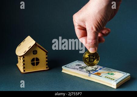 Un homme met une pièce de bitcoin sur une pile de billets de cent dollars devant une maison en bois de jouet sur un fond gris. Banque D'Images