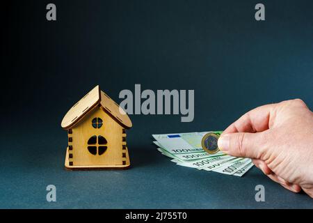 Homme mettant une pièce d'un euro sur une centaine de billets en euros devant une maison en bois sur fond gris, gros plan. Banque D'Images