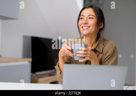 Jeune femme asiatique souriante et gaie prenant un café le matin tout en regardant loin tout en travaillant à l'ordinateur portable, en parlant avec son ami virtuel, assis au bureau à la maison Banque D'Images