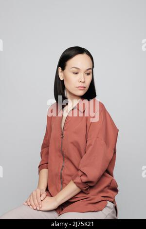 Triste jeune femme asiatique en chemise beige foncé, assise sur un tabouret et regardant vers le bas Banque D'Images