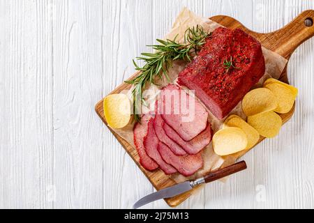 pastrami de bœuf frais coupé en tranches sur une planche en bois avec du romarin frais et des chips de pommes de terre sur une table blanche, vue horizontale depuis le dessus, gros plan Banque D'Images