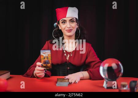 Soulage gitan souriant tenant la carte de tarot près de l'orbe flou isolé sur le noir Banque D'Images