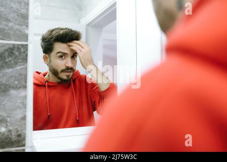 Posé devant le miroir, le jeune homme prend ses mains sur ses cheveux, un homme adulte se préparant à sortir dans la salle de bains Banque D'Images