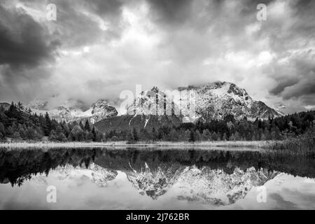 Les montagnes du Karwendel se reflètent dans le lac Luttensee, près de Mittenwald. De la neige fraîche et des nuages atmosphériques encadrent les montagnes. La conversion en noir et blanc donne à la crête de montagne une apparence massive. Banque D'Images