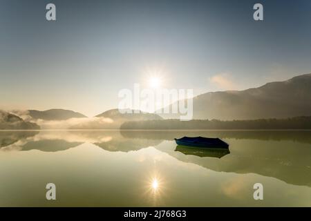 Un seul bateau à rames avec une bâche est ancré au lac Sylvensteinspeicher, tandis que le soleil envoie ses rayons au-dessus du lac avec une légère brume en début de matinée, se reflétant dans l'eau. Banque D'Images