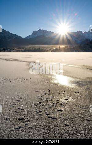 Une étoile du soleil se levant sur les montagnes Karwendel sur la rive du lac Barmsee avec de la glace en premier plan et les montagnes en arrière-plan sous un ciel bleu en hiver. Banque D'Images