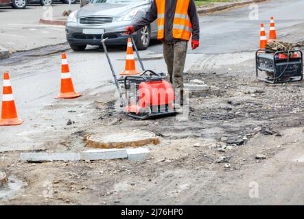 Un ouvrier de service routier bélier une section de la chaussée clôturée avec des cônes de trafic orange près des trous d'égout avec un rameur d'essence. Banque D'Images