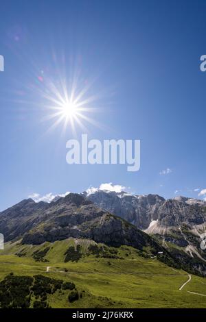 Le célèbre Karwendelhaus dans le Karwendel, dans les Alpes tyroliennes sous le ciel bleu et le soleil avec une étoile du soleil. Au premier plan, les pâturages du Graßlegerbichl, au milieu du sol dominé par la montagne locale de la hutte, le Hochalmkreuz (2192m) et en arrière-plan Schlauchkakopf (2500m), Birkkarspitze (2749m) et l'Ödkarspitzen. Banque D'Images