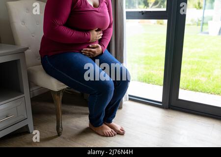 Basse section de la femme afro-américaine moyenne adulte souffrant de maux d'estomac tout en étant assise sur une chaise Banque D'Images