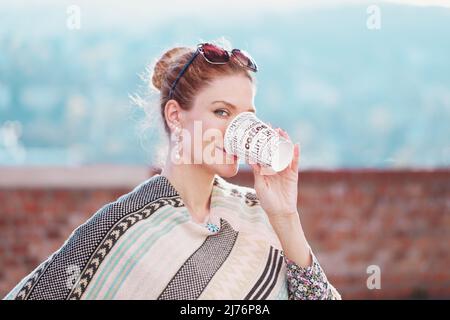 Jeune femme joyeuse à tête rouge dans poncho boire du café à l'extérieur, regardant dans l'appareil photo Banque D'Images