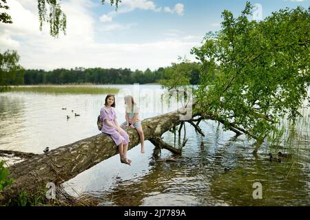 Deux jeunes filles mignonnes assises sur un arbre de faller au bord de la rivière ou du lac en plongeant leurs pieds dans l'eau le chaud jour d'été. Activités familiales en été. Banque D'Images