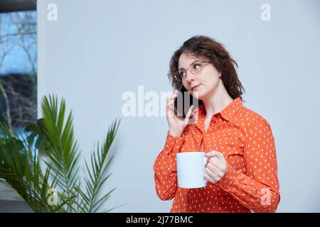 Une femme d'âge moyen parle au téléphone à la maison. Contient le gobelet Banque D'Images