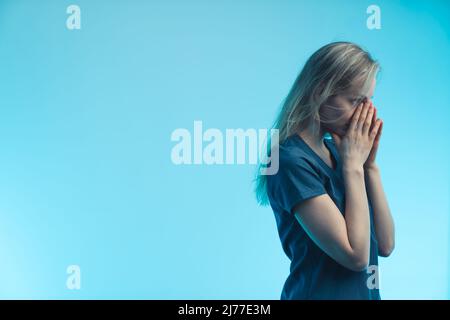 Moyenne studio tourné sur fond bleu d'une femme blonde européenne inquiète médecin avec ses mains pliées près de son visage. . Photo de haute qualité Banque D'Images