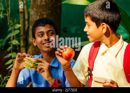 un garçon d'école indien mangeant un sandwich et son ami avec un sac à dos est assis sur une table et mangeant une pomme dans le parc Banque D'Images