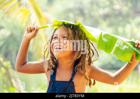 Mowgli garçon indien avec des dreadlocks cheveux se cachant tenant la banane leav comme une couverture de pluie dans les tropiques vert forêt fond Banque D'Images