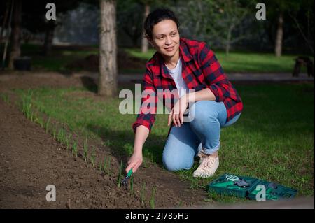 Joyeux charmant jardinier féminin travaille sur un jardin de campagne, libère le sol noir avec des instruments de jardinage et des sourires regardant de côté. H Banque D'Images