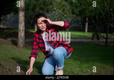 Agréable sereine femme d'âge moyen dans une chemise à carreaux, en regardant de côté sourire, couvrant son visage avec sa main de la lumière du soleil travaillant sur le noir Banque D'Images