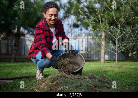 Afro-américaine endurctrice femme au foyer jardinier en gants de travail collectant l'herbe fauchée dans un seau assis près des fourches allongées d Banque D'Images