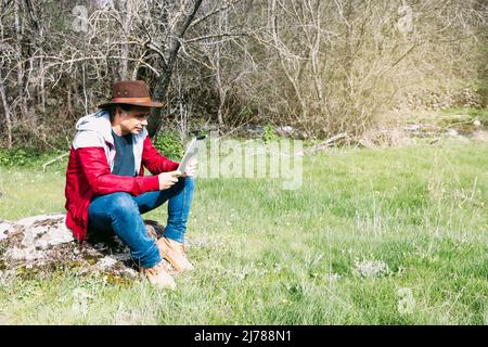 Homme d'entreprise indépendant portant un chapeau, regardant sa tablette tout en se relaxant sur le terrain, assis sur une pierre. Concept de travail, profiter, se détendre, moi Banque D'Images