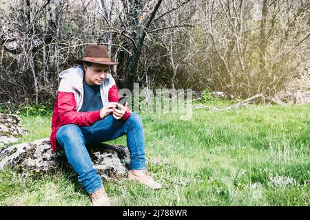 Homme d'affaires indépendant portant un chapeau, regardant son mobile tout en se relaxant dans la campagne, assis sur une pierre. Concept de travail, apprécier, re Banque D'Images