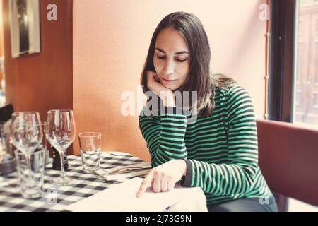 Une jolie jeune dame s'est assise à table pour lire le menu dans un restaurant de bistro tout seul Banque D'Images
